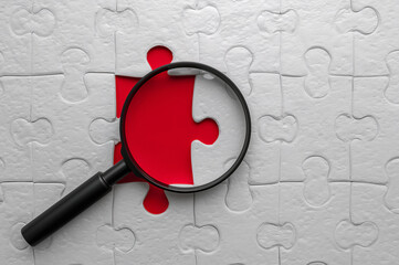 Magnifying glass over a missing red puzzle piece within a white jigsaw puzzle missing piece search