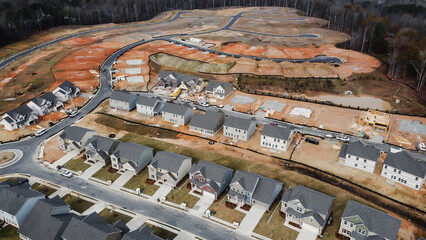 Rows of completed homes with driveways and vehicles, slab foundation lots and timber-framed houses in various stages of construction and site prep, heavy machines working, Flowery Branch, Georgia