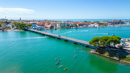 Aerial view of kayaks and boats entering Grado, Italy, with turquoise Adriatic waters, a scenic bridge, and the historic seaside town in the background. Popular destination for water sports, summer 