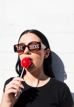 Woman wearing sunglasses with "Life Sucks" inscription, licking a lollipop.