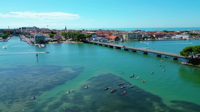 Aerial view of kayaks and boats entering Grado, Italy, with turquoise Adriatic waters, a scenic bridge, and the historic seaside town in the background. Popular destination for water sports, summer 