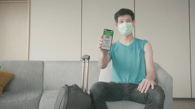 A young man wearing a mask holds a smartphone, showcasing a travel app. He sits on a couch with luggage, highlighting the theme of travel and preparation in a modern context. - Powered by Adobe