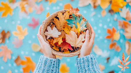 Warm autumn bounty: Hands in soft blue sweater gently hold woven basket brimming with colorful collection of fall leaves, set against cheerful light blue background adorned with scattered foliage.
