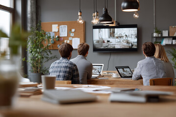 Four people focus on a large screen as they collaborate in a contemporary office setting. Laptops are open, and various work materials are scattered on the table