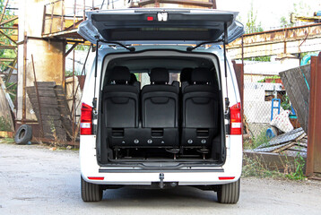 Mini van open trunk. Passenger van trunk. Rear view of white minivan with trunk door open, showing black passenger seats in upright position. Vehicle is parked outdoors near rusty industrial structure