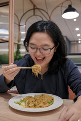Happy woman enjoys stir fried vermicelli noodles with chopsticks in a cozy modern restaurant. The scene captures joyful dining, Asian cuisine.