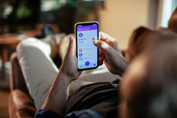 Young man using a banking app on his smart phone while lying down on a couch in the living room