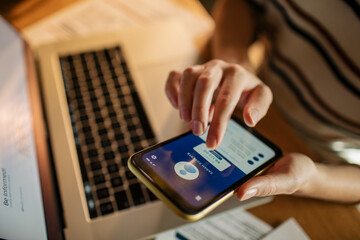 Young woman using a banking app on her smart phone while working on a laptop from home in the kitchen