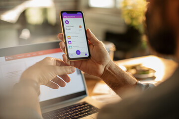 Young man using a banking app on his smart phone while working from home on his laptop in the kitchen