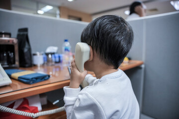 Asian boy is holding a landline phone while sitting at a desk in an office cubicle.