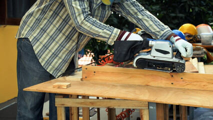 Carpenter sanding wooden board with electric belt sander while wearing protective gloves and striped shirt