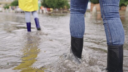 girls in raincoats and rubber boots walk along road flooded with torrential rains, their feet walk...