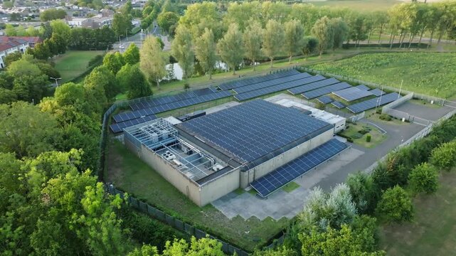 Small modern local data center building in Netherlands with rooftop and ground solar panels surrounded by trees, fence and grassy landscape, aerial orbit