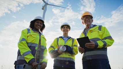 Multi-ethnic group of inspectors finishing discussion and looking directly at camera while smiling. Above wind turbine spinning fast. Proud people. Producing green energy. Saving planet.