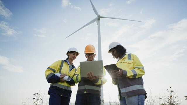 Multi-ethnic group of coworkers standing below tall wind turbine. Looking at laptop screen while comparing data from their architectural plan. Searching for ways of improvement. Personal protection.