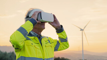 Camera view of Caucasian engineer using 3D glasses for planning new project. Man with yellow costume standing at field. Amazing landscape in background. Sunset. Wind rotating large wind turbine.