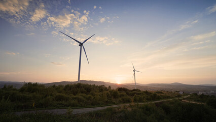 Obraz premium Beautiful view of two wind turbines slowly rotating on top of hill. Blue sky. Bright sunset visible on horizon. Green energy production concept. Caring about nature. Healthy planet.