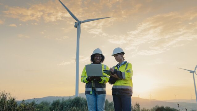 Twi multi-ethnic women working together next to large wind turbines. Wearing helmets and yellow jackets. Coworkers discussing about improving production of green energy. Using portative laptop.