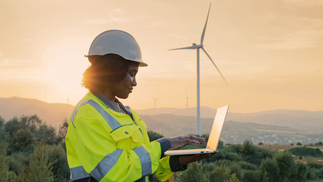 Side view of African American woman working at wind power station. Wearing yellow protective jacket with gray-green inserts and safety helmet. Using laptop for productive work. Renewable green energy.
