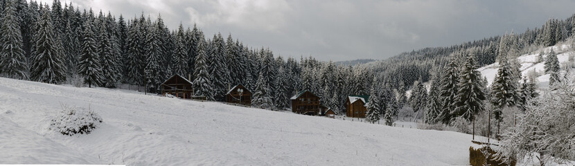 Naklejka premium Small wooden chalets peek through the snow-covered trees, evoking warmth and coziness. The cloudy sky adds a soft, tranquil light, emphasizing the fairy-tale charm of the winter 