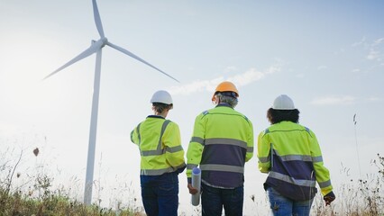 View from behind of multi-ethic workers walking towards spinning wind turbine. Pointing at working mechanism. Wearing personal protective equipment. Blue sky. Inspecting power plant. Engineers.