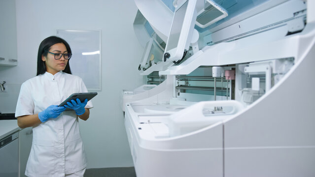 Asian woman medical technician in a lab coat uses a tablet to monitor robotic laboratory equipment, ensuring precise operations.