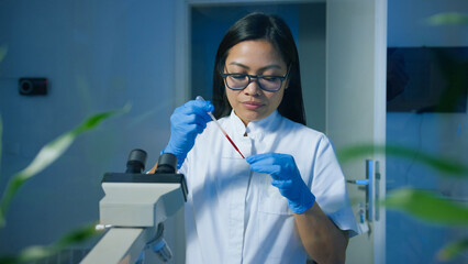 Woman researcher in a lab wears gloves and glasses while analyzing samples with a microscope under dim lighting. © 24K-Production