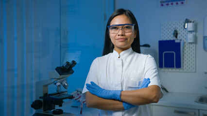A confident asian female scientist stands in a laboratory, arms crossed, wearing protective gear and smiling at the viewer. © 24K-Production