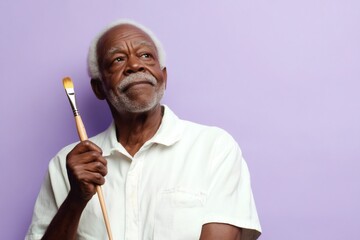 Elderly African American artist contemplates his next stroke, holding a paintbrush and looking upwards on a vibrant purple background