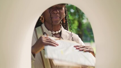 African American female mail carrier wearing cap and crossbody bag delivering envelopes into mailbox while standing in sunlit park