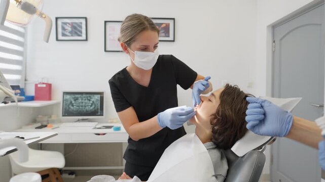 A pediatric dentist applies a topical anesthetic gel to a boy's gums with a cotton swab. This numbing procedure prepares the patient for a painless milk tooth extraction in the dental clinic.