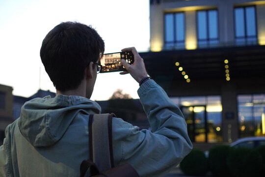  Individual takes a photo of a modern building with a smartphone, reflecting urban exploration. The perspective from behind highlights architecture and technology engagement.