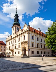 Fototapeta premium A magnificent, light-beige historical building, showcasing a tall, dark-green turret, stands proudly in a sun-drenched square, with a statue at its base, and mature trees casting a gentle shade.