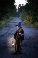 Caucasian blonde child girl in witch black costume with broom and lantern, in outdoor forest background. Vertical shot.