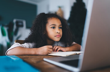 Thoughtful cute black girl doing home work while sitting at table with laptop