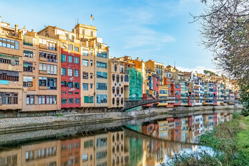 Colorful houses of Girona reflect in the Onyar River at Pont d'en Gomez Bridge, Spain. Vibrant yellow, red, orange, and blue facades create a picturesque scene under clear blue skies