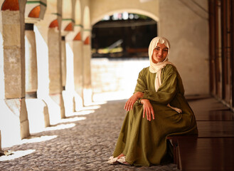 Smiling woman in green modest dress and white hijab sitting outdoors under historic arches, symbolizing elegance, culture, tradition and modern lifestyle.