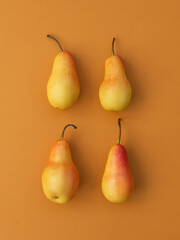 Four ripe yellow and red pears arranged in two rows on a muted orange background with soft shadows and natural light