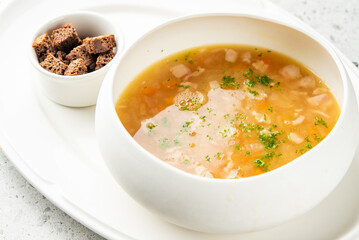 Clear broth soup with meat, vegetables, and herbs served in a white bowl, accompanied by croutons. Minimalist food presentation on a white plate, top view.