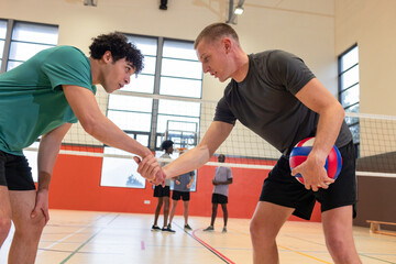 Male volleyball players shaking hands at net in school gym near bench, ball, court markings