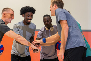 Diverse male teammates stacking hands in sports hall holding volleyballs, wearing headphones