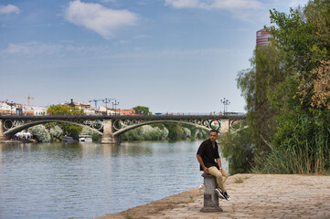 Fototapeta premium Young, handsome man with a beard, wearing a black shirt and beige cargo trousers, sitting on a metal poster used to moor boats,with the river and the famous Triana Bridge in Seville in the background.