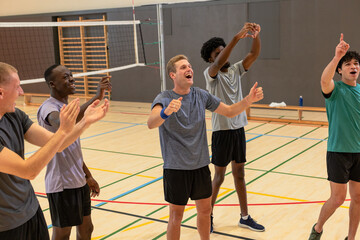 Diverse male volleyball team clapping, raising arms on gym court under net with water bottle