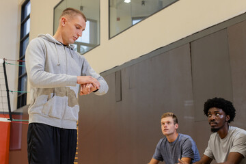 Diverse volleyball coach checking wristwatch and players kneeling beside net on floor lines