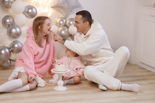 Young Caucasian parents in hoodies with baby girl in pink outfit, sitting on wooden floor, balloons and cake, playful moment, birthday party at home. Concept: family, love, birthday, celebration.