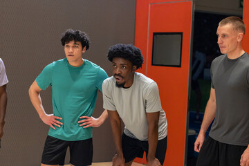 Diverse male teammates standing on wooden basketball court near padded grey wall, orange door