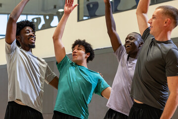 Diverse male friends in sportswear raising arms celebrating on basketball court with padded walls