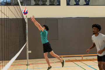Male volleyball players in teal, grey shirts blocking red-white-blue volleyball at net in gym