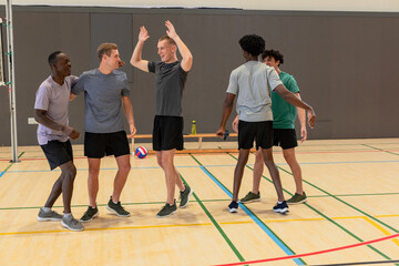 Diverse male teammates in sports wear high-fiving while celebrating on gym court with volleyball