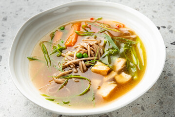 Bowl of soba noodle soup with tofu, carrots, peas, and leafy greens in clear broth. Healthy Asian meal, top view on light background. Natural light, vibrant colors